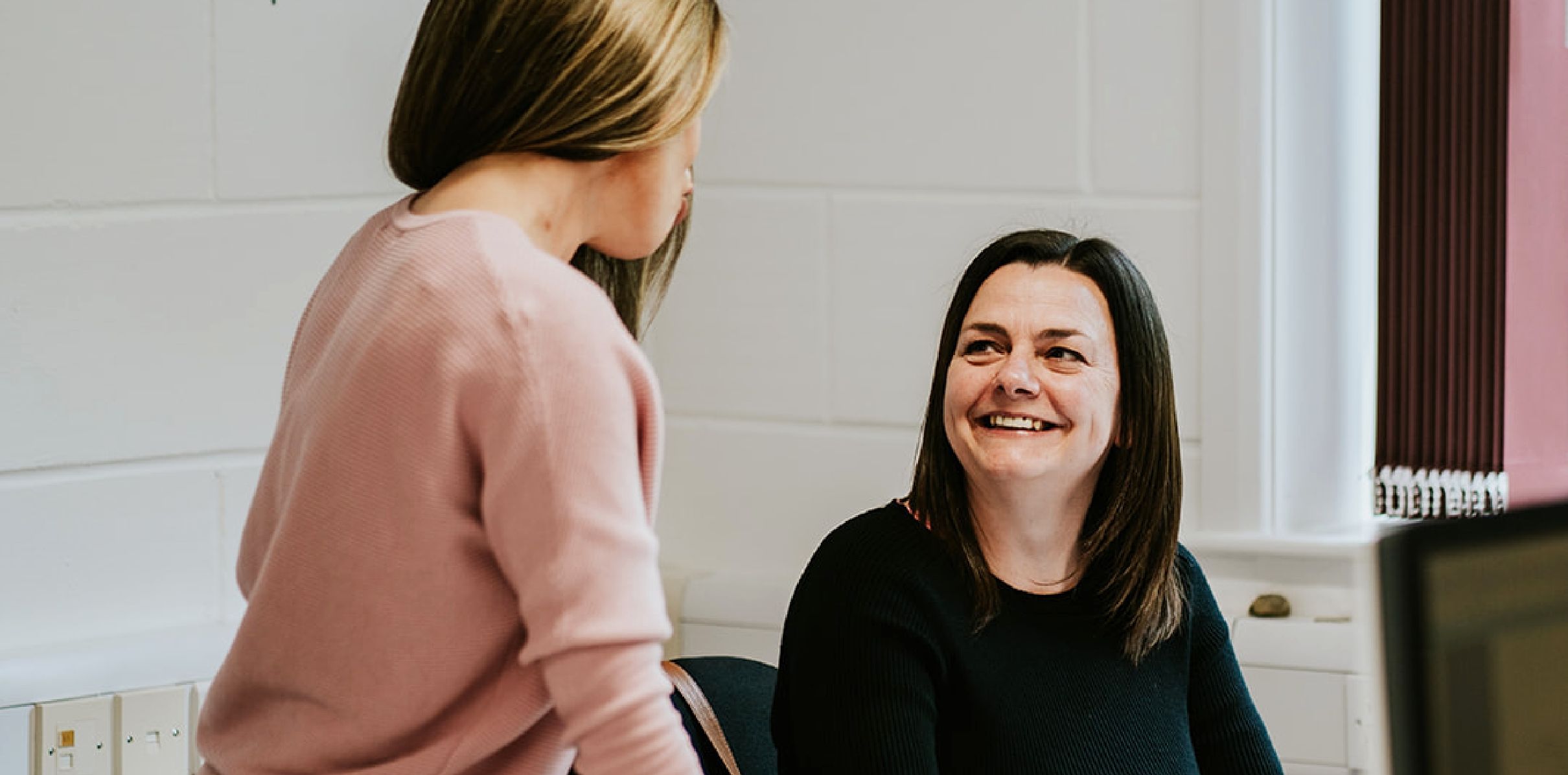 A woman, sat down, smiling at another woman, standing up