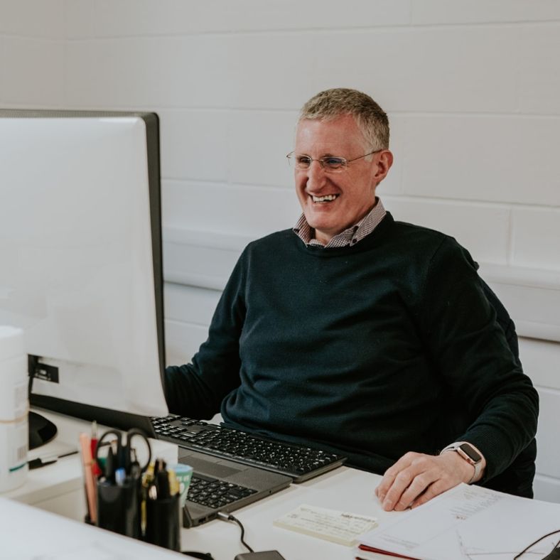 A man sat behind a desk smiling at a computer monitor